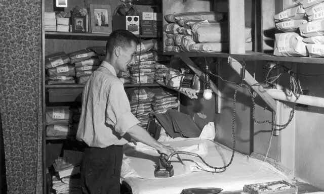 Um trabalhador de lavanderia passa uma camisa com prateleiras ao seu redor segurando roupas limpas embrulhadas em papel pardo, por volta de 1945. (Lawrence Thornton/Getty Images)