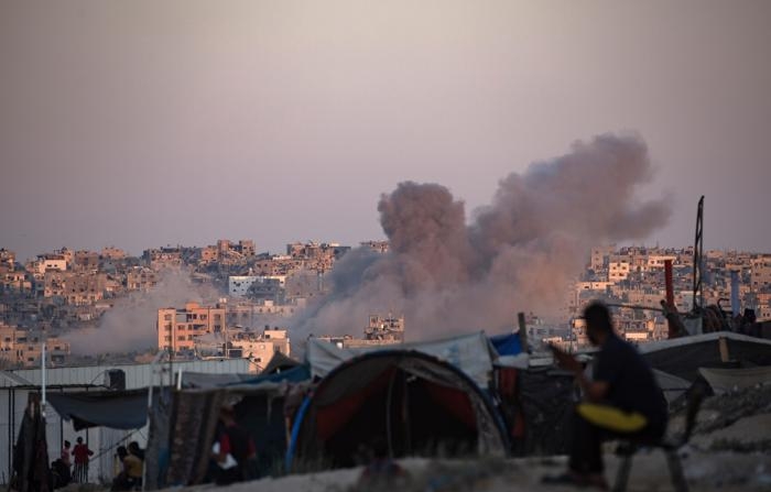 A fumaça sobe após um ataque aéreo israelense enquanto palestinos deslocados internamente sentam-se ao lado de suas tendas no campo de Khan Younis, no sul da Faixa de Gaza (EFE/Haitram Imad).