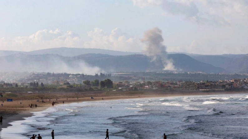 Pessoas caminham em uma praia enquanto a fumaça surge ao fundo no lado libanês da fronteira com Israel, vista de Tiro, no sul do Líbano, em 22 de setembro de 2024 (Aziz Taher/Reuters)