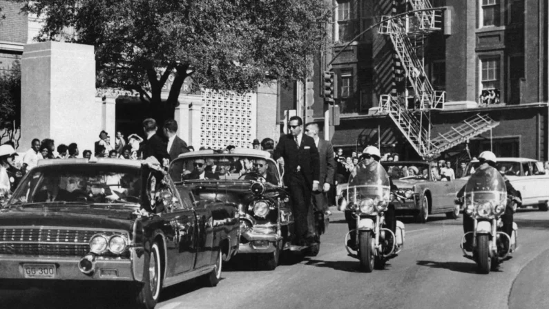 Vista através do para-brisa do conversível em primeiro plano, a mão do presidente John F. Kennedy alcança sua cabeça segundos depois de ser morto a tiros, enquanto a primeira-dama Jacqueline Kennedy segura seu antebraço enquanto a carreata segue pela Elm Street, passando pelo Texas School Book Depository, em Dallas, em 22 de novembro de 1963 (Foto de James W. "Ike" Altgens / AP)
